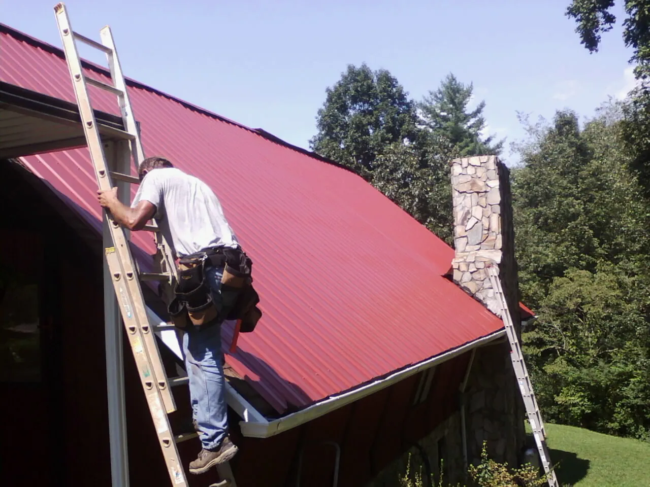 Roofer on a ladder beside a red metal roof and stone chimney — Soffit & Fascia Repair by Roofing Brothers in Islandton